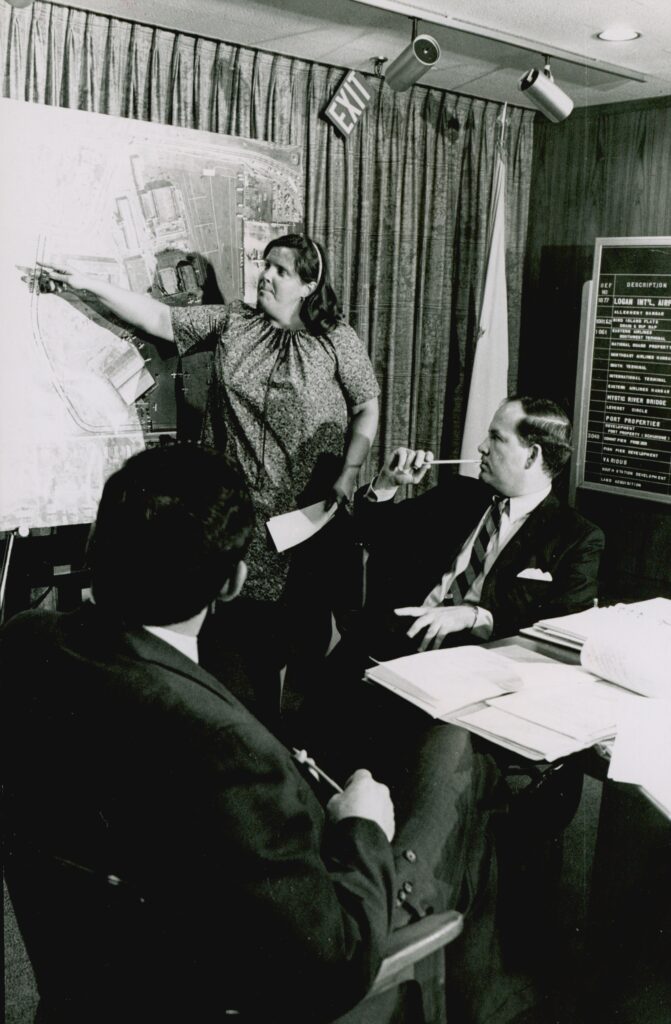黑白的照片,一个女人站在城市规划的地图和指向,而两个坐着的人看。