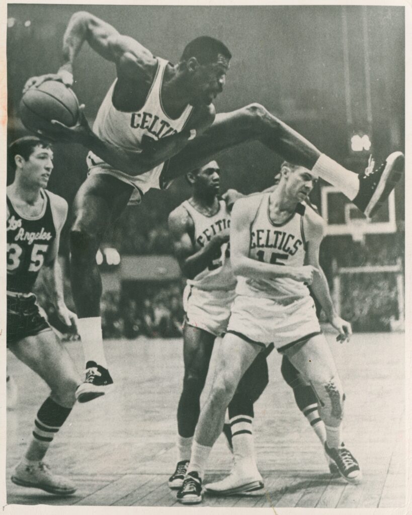 黑白照片中，身穿白色凯尔特人队服的比尔·拉塞尔双手拿着球，两腿分开跳跃。