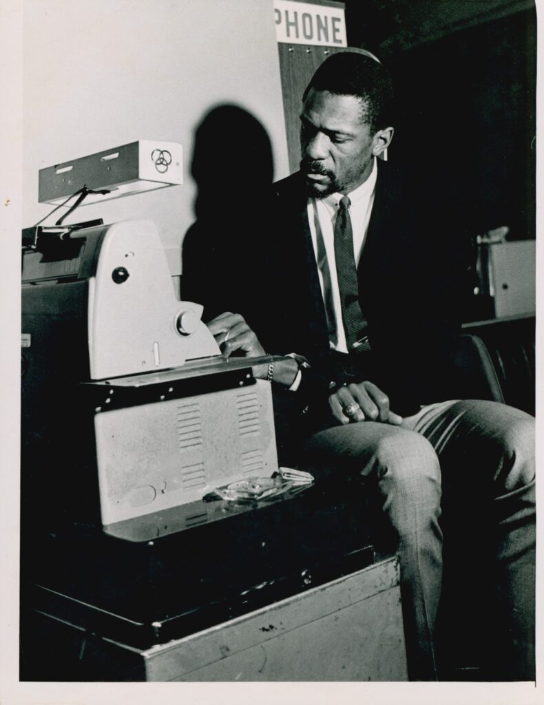比尔·拉塞尔穿着深色夹克，打着领带，坐在一台机器前的黑白照片。
