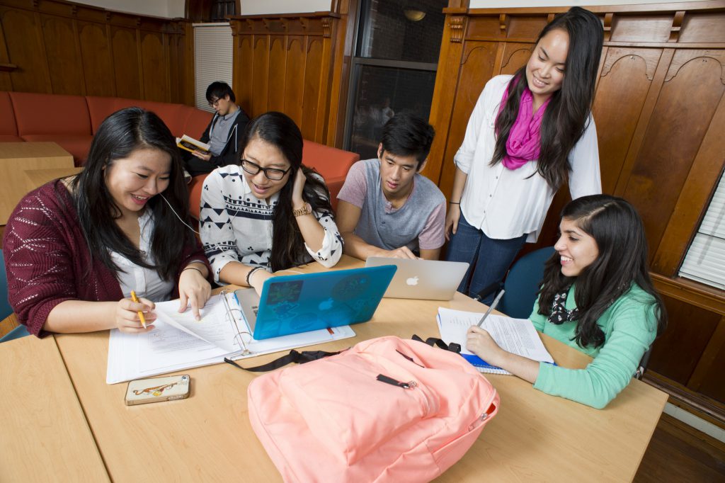 学生围坐在一张桌子的形象。他们正在写论文,一个学生有自己的笔记本电脑打开。